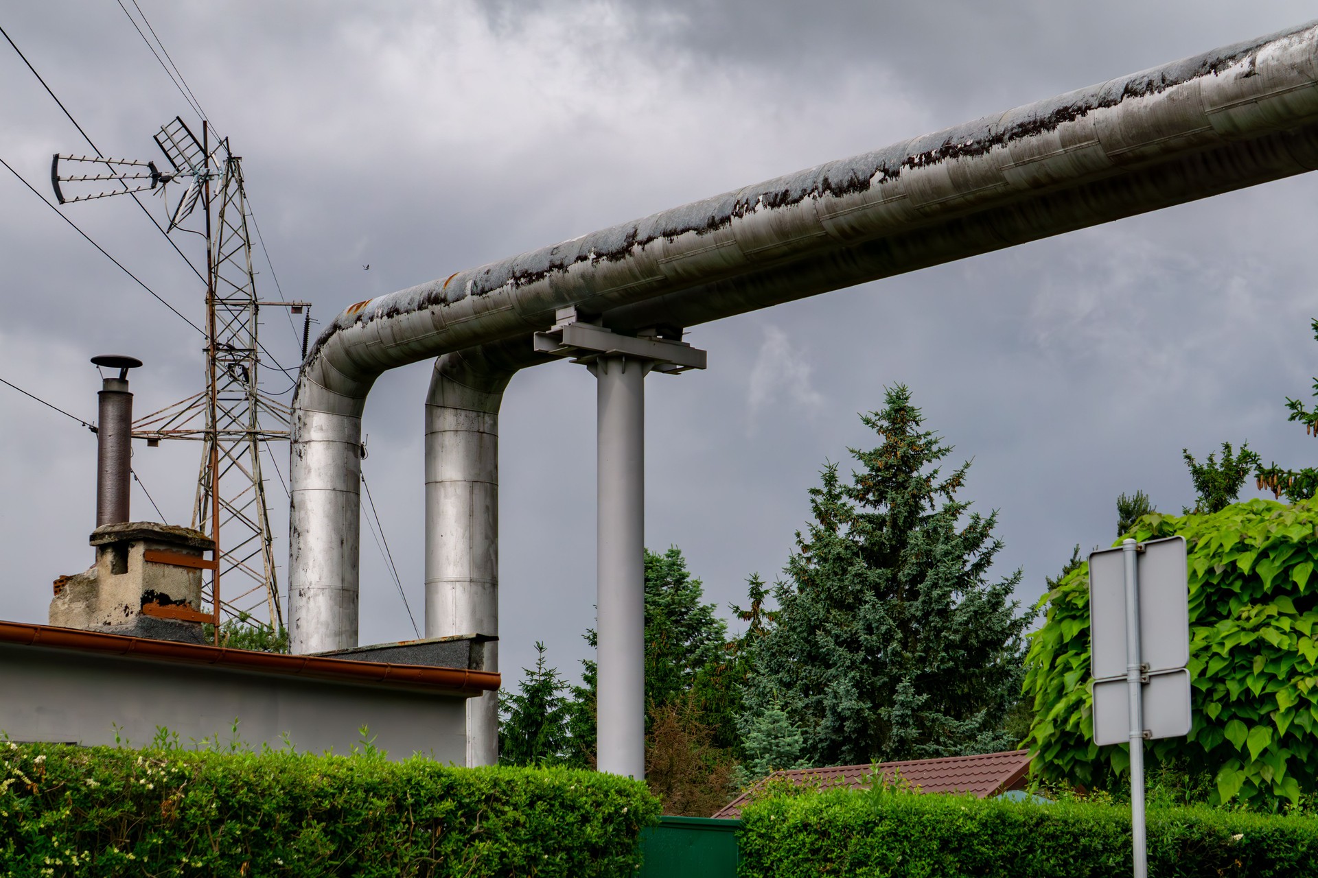 Tuyaux de chauffage aériens au-dessus d’une rue, soutenus par des structures en acier. À l’arrière-plan, des bâtiments industriels, des cheminées et un poteau électrique, le tout sous un ciel sombre et nuageux